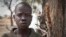 In this photo taken Saturday, Feb. 15, 2014, Ajing Abiik, 12, who fled from his hometown of Bor when the fighting broke out in December, stands next to the tree where he and his two brothers sleep in Minkaman IDP camp, Awerial County, in South Sudan. The fighting in the world’s newest country has left thousands of its youngest citizens either orphans or separated from their parents, increasing their vulnerability to sickness, malnutrition and recruitment by warring groups as child soldiers. (AP Photo/Mackenzie Knowles-Coursin)