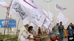 Sejumlah warga berada di pinggir jalan yang dihiasi bendera Taliban di Kabul, Afghanistan, pada 15 Agustus 2023. Bendera tersebut dipasang sebagai perayaan dua tahun kembalinya Taliban berkuasa di negara itu. (Foto: AP/Abdul Khaliq)