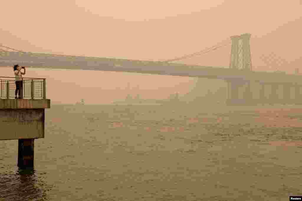 A person wearing a face mask takes a picture of the Manhattan skyline while it&#39;s covered in haze and smoke caused by wildfires in Canada, in Brooklyn, New York, June 7, 2023.