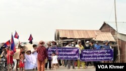 រូបឯកសារ៖ ពលរដ្ឋរស់នៅក្នុងសង្កាត់សំរោងត្បូងជាប់ផ្ទៃបឹងតាមោក ឈរតវ៉ាលើកបដានៅមុខផ្លូវទំនប់កប់ស្រូវនាពីថ្ងៃទី២៧ ខែកុម្ភៈ ឆ្នាំ២០២៤។ (ស៊ឹម ចាន់សំណាង/វីអូអេ)
