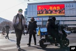 Orang-orang berjalan di seberang jalan dekat layar besar yang mempromosikan Tentara Pembebasan Rakyat China dengan tulisan "Berdarah" di luar sebuah mal di Beijing, Senin, 9 Januari 2023. (Foto: AP)