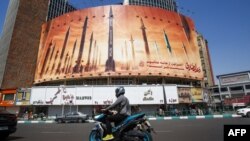 បុរសម្នាក់ចុះម៉ូតូកាត់ផ្ទាំងផ្សាយពាណិជ្ជកម្មដែល​បង្ហាញពី​មីស៊ីលអ៊ីរ៉ង់នៅតេហេរ៉ង់នៅថ្ងៃទី១៩ ខែមេសា ឆ្នាំ២០២៤។ (AFP)