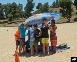 Youths shelter in the shade after being asked by lifeguards to retrieve their footwear before getting onto the sunny beach area to prevent burned feet at Castaic Lake beach as the temperature rises, July 8, 2024, in Castaic, Calif.