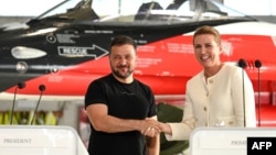 ปธน.โวโลดิเมียร์ เซเลนสกี แห่งยูเครน (ซ้าย) และนายกรัฐมนตรีเมตเต เฟรเดริกเซน แห่งเดนมาร์ก (ขวา) จับมือกันระหว่างการแถลงข่าวร่วมที่หน้าเครื่องบินเอฟ-16 ในโรงจอดเครื่องบินของฐานทัพอากาศในเมืองโวเจนส์ ทางเหนือของเดนมาร์ก เมื่อวันที่ 20 ส.ค. 2023