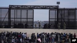 FILE - Migrants who crossed the border from Mexico into the US wait next to the US border wall where US Border Patrol agents stand guard, seen from Ciudad Juarez, Mexico, March 30, 2023. 