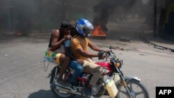 A woman with a child lowers her head as she leaves the Port-au-Prince area on a motorcycle, March 20, 2024. Amid gunfire and a shortage of resources, humanitarians are trying to help as many of the 5.5 million Haitians in need as they can.