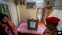 Seorang petugas keamanan berjaga ketika Rubi Paul, 89 tahun, duduk di tempat tidurnya di belakang layar pemungutan suara portabel, memberikan suaranya di kediamannya di Guwahati, India, Minggu, 28 April 2024. (Foto: AP)