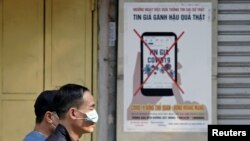 បុរស​ពីរ​នាក់​ដើរ​កាត់​ផ្ទាំង​បដា​មួយ​ដែល​ព្រមាន​ពី​ការ​ចែកចាយ​ព័ត៌មាន​ប្រឌិត​លើ​អ៊ីនធឺណិត​អំពី​ជំងឺ​កូវីដ១៩ ទីក្រុង​ហាណូយ ប្រទេស​វៀតណាម ថ្ងៃទី១៤ ខែមេសា ឆ្នាំ២០២០។ 