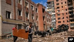 Two men carry a wood panel next to damaged buildings and destroyed cars in a Russian bombing in Bakhmut, eastern Ukraine, May 24, 2022. The town of has been coming under increasing artillery strikes over the last week as Russian forces try to encircle the city of Sieverodonetsk.