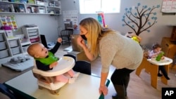 FILE - Amy McCoy signs to a baby about food as a toddler finishes lunch behind at her Forever Young Daycare facility, Monday, Oct. 25, 2021. (AP Photo/Elaine Thompson, File)
