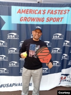 Tarmizi Mawardi memegang bola dan paddle, berfoto di depan backdrop bertuliskan "Olahraga Paling Pesat di AS"