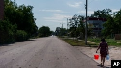 A resident carries water from a well down a road damaged by shrapnel from a May rocket attack in Sloviansk, Donetsk region, eastern Ukraine, Saturday, Aug. 6, 2022. (AP Photo/David Goldman) 