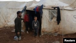 បុរស​ម្នាក់​ឈរ​នៅ​ជិត​កូនរបស់​លោក​នៅ​ខាង​ក្រៅ​តង់​មួយ​នៅ​ជំរំ​ជន​ផ្លាស់ប្ដូរ​លំនៅ​ក្នុង​ស្រុក​ នៅ​ភាគខាងជើង​ខេត្ត Aleppo ក្បែរ​ព្រំប្រទល់​រវាង​ប្រទេស​ស៊ីរី និង​ប្រទេស​តួកគី ថ្ងៃទី១៧ ខែកុម្ភៈ ឆ្នាំ២០២១។