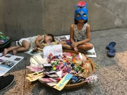 A young girl sits next to a sleeping baby as she sells accessories on the sidewalk in Bangkok, Thailand.