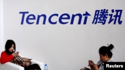 Dua orang pengunjung sedang bermain dengan telepon genggam mereka, duduk di bawah logo perusahaan raksasa internet Tencent, saat berlangsungnya Konferensi Global Mobile Internet di Beijing, 6 Mei 2014. (Foto: dok).