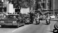 FILE - Seen through the foreground convertible's windshield, President John F. Kennedy's hand reaches toward his head within seconds of being fatally shot as first lady Jacqueline Kennedy holds his forearm in Dallas, on Nov. 22, 1963.