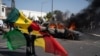 A demonstrator holds a Senegalese flag as he kneels in front of a burning car during protests against the arrest of opposition leader and former presidential candidate Ousmane Sonko, Senegal, March 8, 2021.