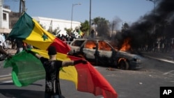 A demonstrator holds a Senegalese flag as he kneels in front of a burning car during protests against the arrest of opposition leader and former presidential candidate Ousmane Sonko, Senegal, March 8, 2021.