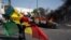 A demonstrator holds a Senegalese flag as he kneels in front of a burning car during protests against the arrest of opposition leader and former presidential candidate Ousmane Sonko, Senegal, March 8, 2021.