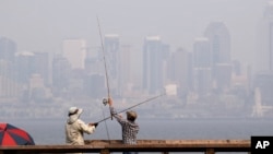A pair of anglers uncross their lines while fishing in Elliott Bay as a smoky haze obscures downtown Seattle in the background, Aug. 14, 2018. Public-health officials Wednesday warned of unhealthy air across many parts of the Pacific Northwest as wildfires send thick smoke across the region, prompting air quality alerts.
