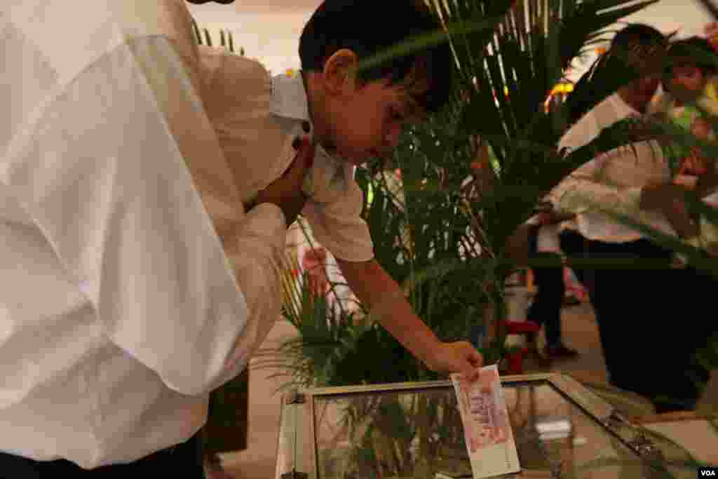 ពុទ្ធសាសនិក​មួយ​រូប​លើក​កូន​របស់​ខ្លួន​ដើម្បី​ដាក់​បច្ច័យ​ក្នុង​ហិប​នៅ​ថ្ងៃបុណ្យ​ភ្ជុំបិណ្ឌ ក្នុង​វត្ត​មួយ​កណ្តាល​រាជធានី​ភ្នំពេញ​ កាលពី​ថ្ងៃទី១ ខែតុលា ឆ្នាំ២០១៦។ (ហ៊ាន សុជាតា/VOA)