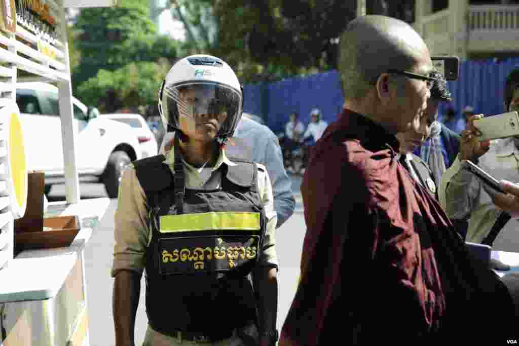 កងកម្លាំងសន្តិសុខមួយរូបឈរយាមកាម ខណៈព្រះសង្ឃសកម្មជនមួយអង្គកំពុងផ្ដល់បទសម្ភាសដល់អ្នកសារព័ត៌មាន អំឡុងពិធីខួបរំឭកវិញ្ញាណក្ខ័ន្ធលោក កែម ឡី នៅស្ថានីយ៍ប្រេងឥន្ធនៈកាល់តិច នៅស្តុបបូកគោ ខណ្ឌចម្ការមន រាជធានីភ្នំពេញ កាលពីព្រឹកថ្ងៃ១០ ខែកក្កដា ឆ្នាំ២០១៩។ (អូន ឆេងប៉រ/VO