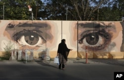 An Afghan woman waits for transportation in front of street art on a barrier wall of the NDS (National Directorate of Security) in Kabul, Aug. 20, 2015.