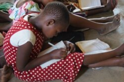 Not an easy task, students sitting on a floor at Mazembe Primary School as a result of poor infrastructure perpetuated by a boom in primary school enrollment. (L. Masina/VOA)