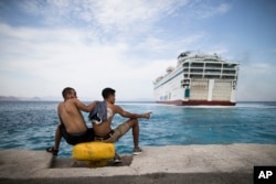 Two migrants look toward a giant passenger ferry while it arrives at the Greek holiday island of Kos, to provide temporary accommodation for some thousands of refugees sleeping rough after crossing clandestinely from Turkey in flimsy boats, Aug. 14, 2015.