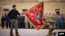 პრეზიდენტი ზელენსკი უკრაინის შეიარაღებული ძალების დროშით ხელში, რომელსაც უკრაინელი ჯარისკაცი პატივს მიაქგებს. 1 ოქტომბერი, 2023. 