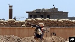 FILE - A Kurdish peshmerga fighter carries his weapon as he walks to his base where two Islamic State flags are seen on a building, right, and a water tower, left, south of Kirkuk, northern Iraq, June 25, 2014. 