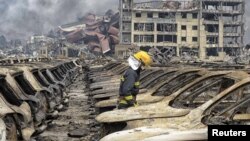 អ្នក​ពន្លត់​អគ្គីភ័យ​ម្នាក់​ដើរ​កាត់​ឡាន​ដែល​ខូច​ នៅ​ពេល​ដែល​ផ្សែង​ហោះហើរ​នៅ​កណ្តាល​កុងតឺន័រ​ដឹក​ទំនិញ​ នៅ​កន្លែង​ផ្ទុះ​ក្នុង​ស្រុក​ថ្មី​មួយ​ដែល​មាន​ឈ្មោះ​ថា Binhai ក្នុង​ក្រុង​ Tianjin ប្រទេស​ចិន កាលពី​ថ្ងៃទី១៤ ខែសីហា ឆ្នាំ២០១៥។