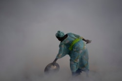 Ndeye Yacine Dieng drops embers over peanut shells covering fish as she walks amidst the smoke on Bargny beach, Senegal, Apr. 21, 2021.