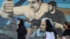 Beberapa perempuan berjalan melewati mural anti-Israel yang menggambarkan pejuang dengan senapan serbu di sepanjang dinding di Alun-Alun Palestina di Tehran pada 14 April 2024. (Foto: AFP)