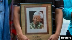 FILE - A man holds a tribute photograph as he watches the funeral service for former South African President Nelson Mandela on a large screen television in Cape Town, December 15, 2013.