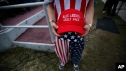 A man dressed in American flag clothes holds a "Make America Great Again" hat before a campaign rally at the Ladd–Peebles Stadium, Dec. 17, 2016, in Mobile, Alabama. Comedians the world over thrive on slogans and this one has not remained unscathed.