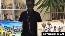 Santo Makoi poses in front of two of his recent paintings of village life in South Sudan. He says that he likes to show people what life is like back home.