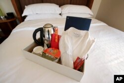 Items in a welcoming kit containing creature comforts that many Chinese travelers expect in their rooms, including, from the left, an electric kettle, green tea, instant noodles, slippers, and a robe, rest in a tray on a bed in Boston.