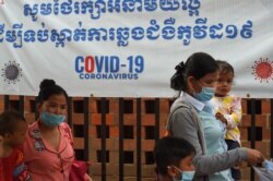 FILE - Women wearing face masks amid the coronavirus pandemic walk with their children outside a hospital with a banner warning about the dangers of the virus, in Phnom Penh, Cambodia, Sept. 29, 2020.