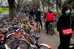 Residents ride bicycles from bike-sharing company Ofo try to pedal through a sidewalk crowded with bicycles from the bike-sharing companies Ofo, Mobike and Bluegogo, near a bus stand in Beijing, China, March 23, 2017.