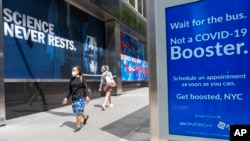 FILE - Pedestrians walk past signs hanging outside Pfizer headquarters in New York and one hanging at a bus stop encouraging the COVID-19 booster, May 23, 2022. 