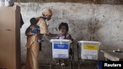 ស្ត្រី⁣ម្នាក់កំពុង⁣ស្ពាយ⁣កូននៅលើខ្នង ដាក់សន្លឹក⁣ឆ្នោត⁣ក្នុងហិប នៅ Freetown ខែវិច្ឆិកា ២០១២។