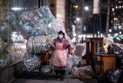 FILE - A woman wearing a face mask and a plastic bag pulls a cart loaded with bags of recyclables through the streets of Lower Manhattan during the outbreak of the novel coronavirus In New York City, April 16, 2020.