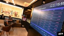 A bank representative and a client wearing facemasks sit near an electronic board displaying global indices price performance at a shopping mall in Bangkok on March 15, 2020.