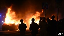 Sejumlah warga tampak melihat kobaran api yang melalap kendaraan di wilayah Rosengard, Malmo, Swedia, menyusul bentrokan yang terjadi pada 4 September 2023. (Foto: Johan Nilsson/TT/TT NEWS AGENCY/AFP)
