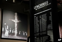 In this Nov. 10, 2017 image, a monolith listing the names, dates and rationale for the lynching of African-American residents rests in the foreground of a photograph of a burning Ku Klux Klan cross on display in the Mississippi Civil Rights Museum.