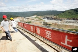 In this Oct. 6, 2012 photo, a Chinese engineer, left, walks by a fence with Chinese slogans reading: Safety first, highest quality and quantity near an entrance of a dam construction site by China National Heavy Machinery Corporation on the Tatay River in Koh Kong province, some 210 kilometers (130 miles) west of Phnom Penh, Cambodia.