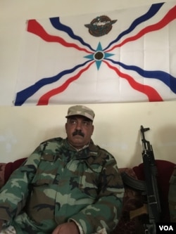 Dwekh Nawsha Col Sameer Oraha sits under the Assyrian Patriotic Party flag, his weapon propped up next to him. (S. Behn/VOA)