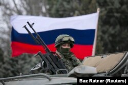 Seorang pria pro-Rusia (tidak terlihat) memegang bendera Rusia di belakang seorang prajurit bersenjata di atas kendaraan tentara Rusia di luar pos penjaga perbatasan Ukraina di kota Balaclava, Krimea. (Foto: REUTERS/Baz Ratner)