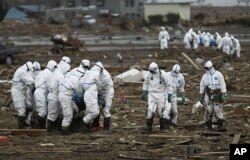 មន្រ្តី​ប៉ូលីស​ជប៉ុន​មួយក្រុម​ពាក់​សម្លៀកបំពាក់​ព៌ណស​ដើម្បីការពារ​សារជាតិ​រ៉េអាក់ទ័រ លើក​ជនរងគ្រោះ​ម្នាក់ ខណៈដែល​មួយក្រុម​ទៀត​លើក​សាកសព​នៅ​ខាងក្រោយ ក្នុង​ពេល​ស្វែងរក​មនុស្ស​ដែល​បាត់ខ្លួន​នៅ Minami Soma នៅ Fukushima Prefecture នៅ​ប្រទេស​ជប៉ុន កាលពីថ្ងៃទី៨